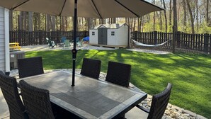 Une terrasse avec une table et des chaises, un parasol, un petit abri de jardin et un hamac.