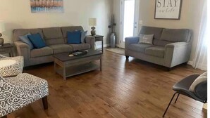 Un salon avec deux canapés gris, un pouf à motifs, une table basse et un tableau encadré accroché au mur.