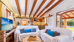A cozy living room with two white sofas, a coffee table with chess and snacks, a TV, and a kitchen in the background.
