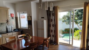 Une cuisine avec des meubles en bois, une table à manger avec des chaises, un réfrigérateur et une vue sur un jardin, visible à travers une porte-fenêtre coulissante.