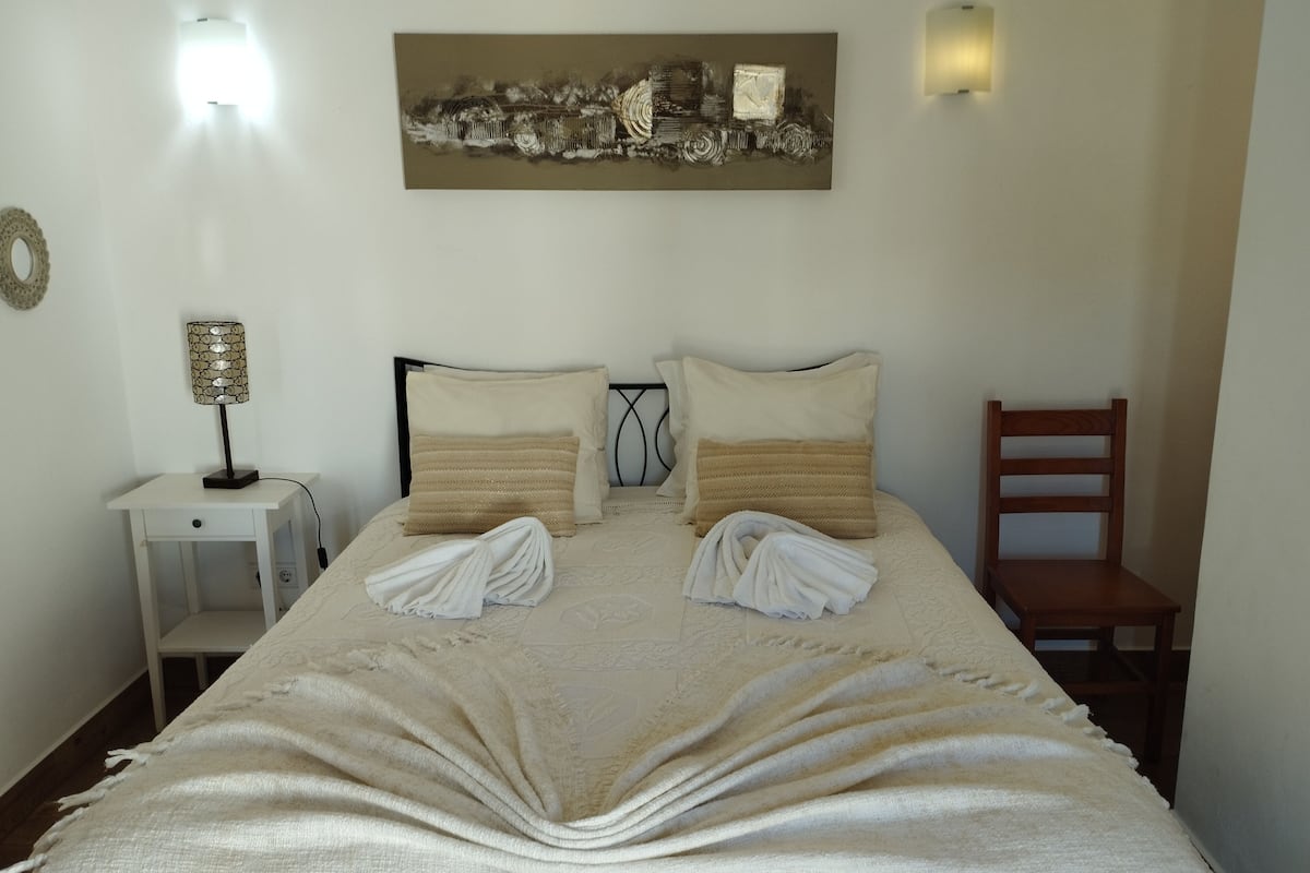 Una cama bien hecha con dos almohadas, una lámpara de noche y un cuadro en la pared.