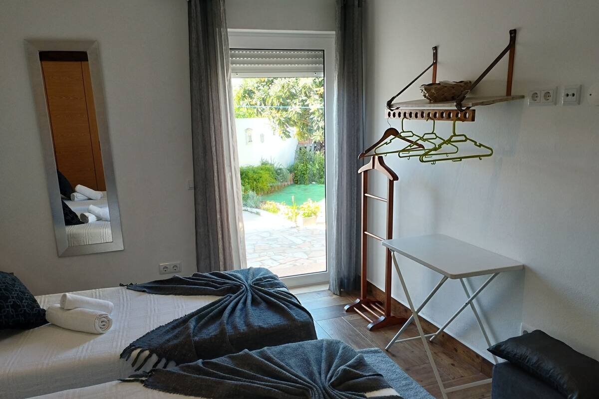 Habitación de hotel con dos camas, una mesita, un espejo y vista a un jardín a través de un ventanal.