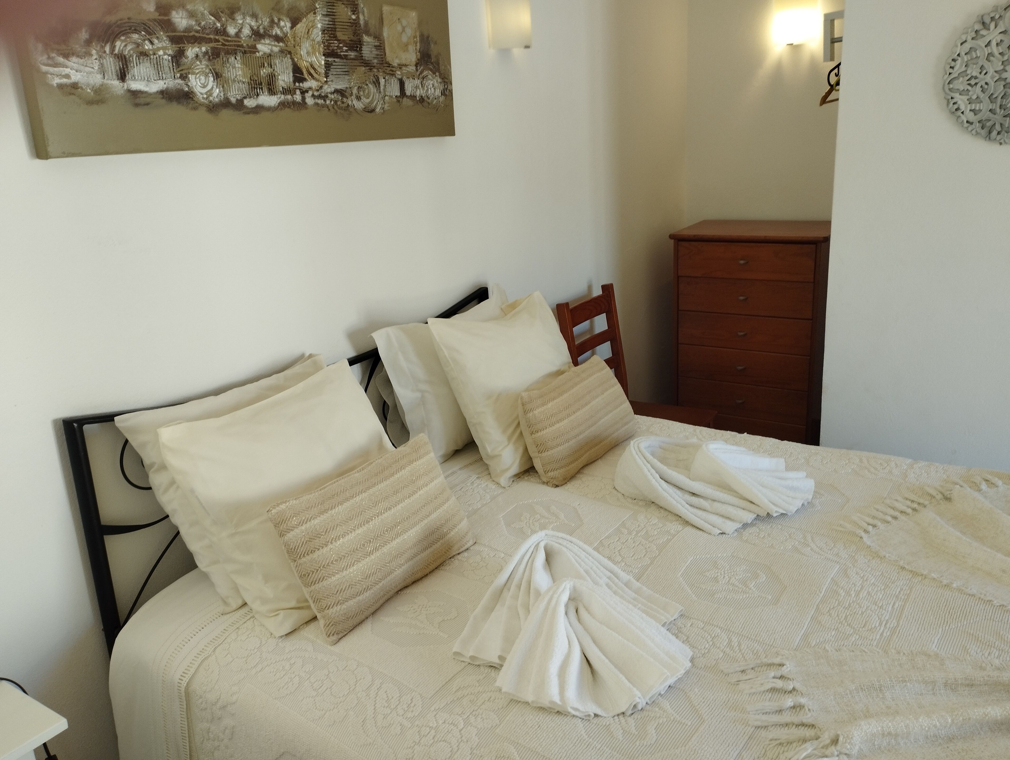 A neatly made bed with pillows and towels, a wooden nightstand, a dresser, and a wall-mounted light fixture.