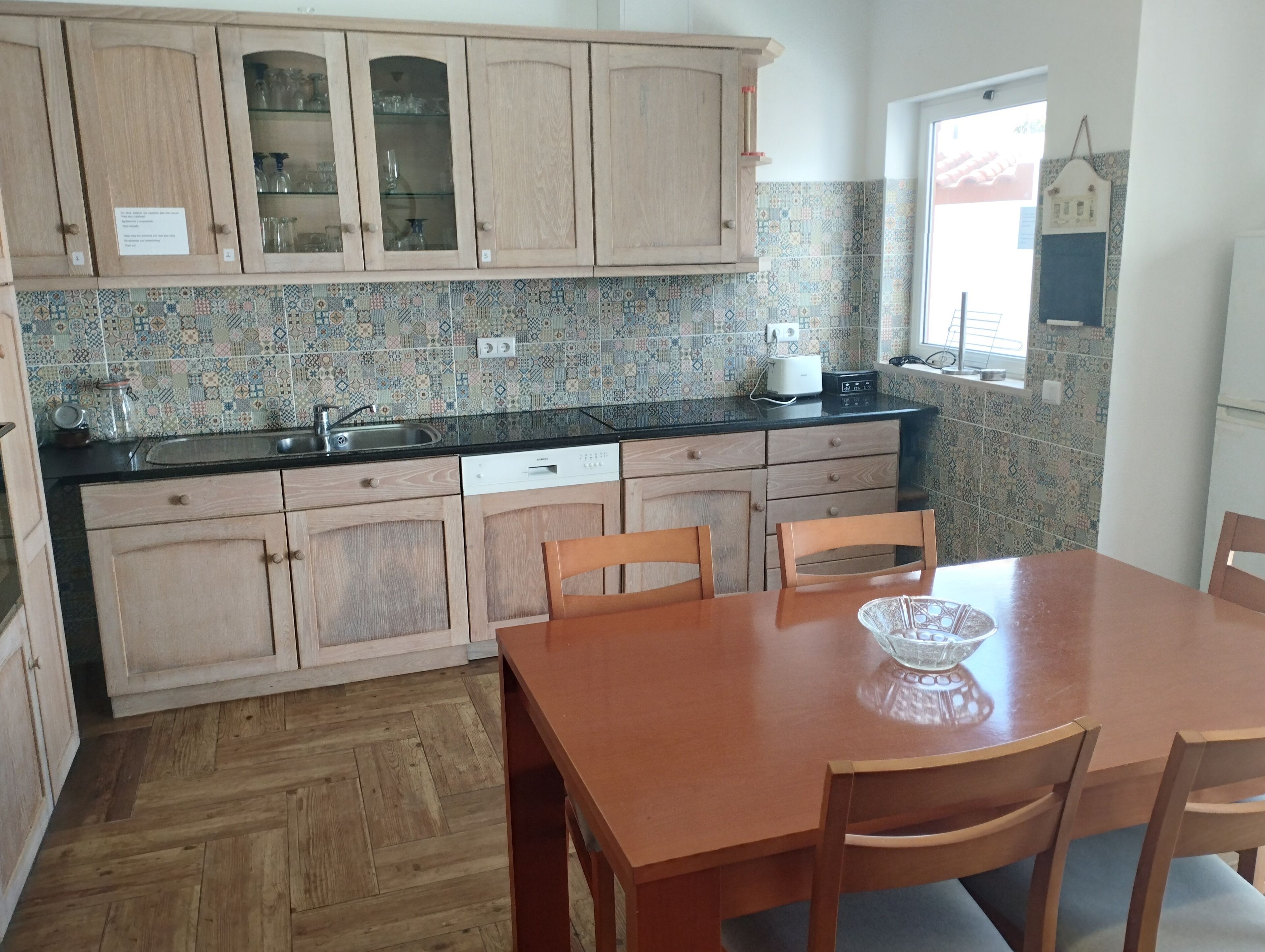 A kitchen with wooden cabinets, a tiled backsplash, a black countertop, a sink, a dishwasher, and a dining table with chairs.