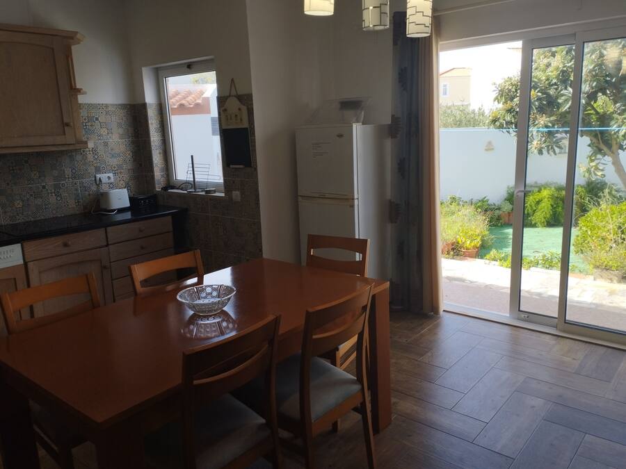Una cocina con mesa y sillas, un refrigerador y ventana con vista a un jardín.