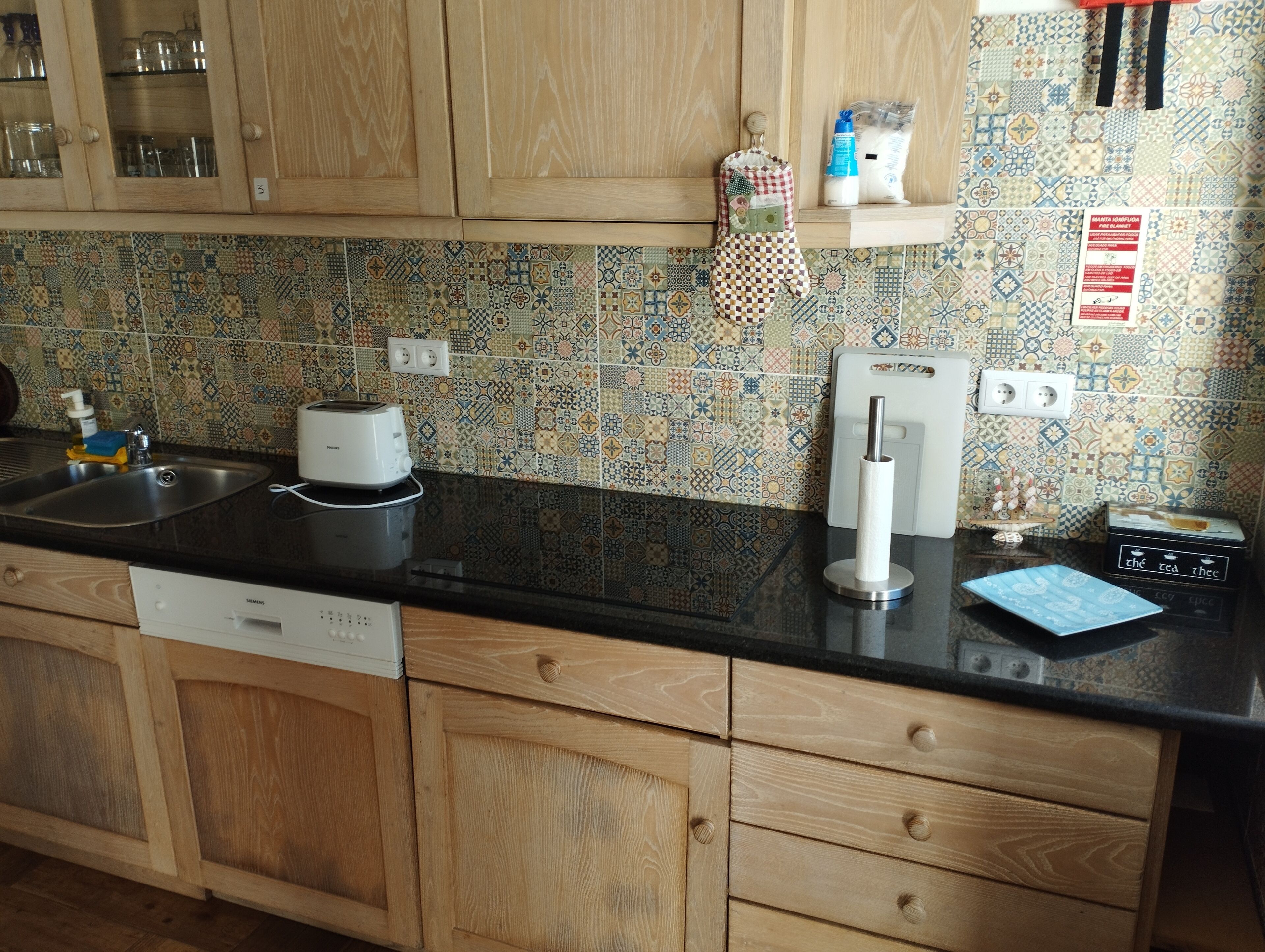 Una cocina con armarios de madera, encimera negra, lavavajillas Siemens, tostadora, dispensador de toallas de papel y un salpicadero decorativo de azulejos.