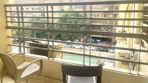 Ein Balkon mit Metallgeländern, zwei Korbstühle und Blick auf eine belebte Straße mit Autos und Gebäuden im Hintergrund.