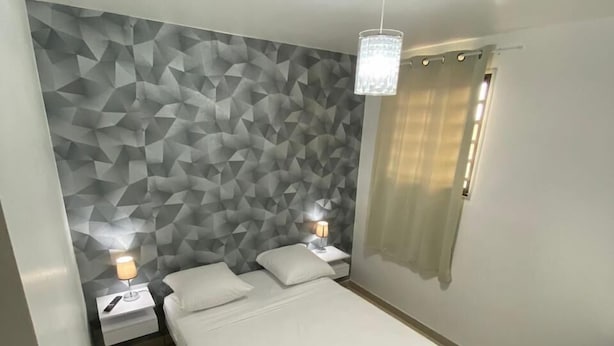 A modern bedroom with a geometric-patterned wall, a bed with white linens, two bedside tables with lamps, and a ceiling light fixture. - LE CHANNEL HOTEL (Le Marin)