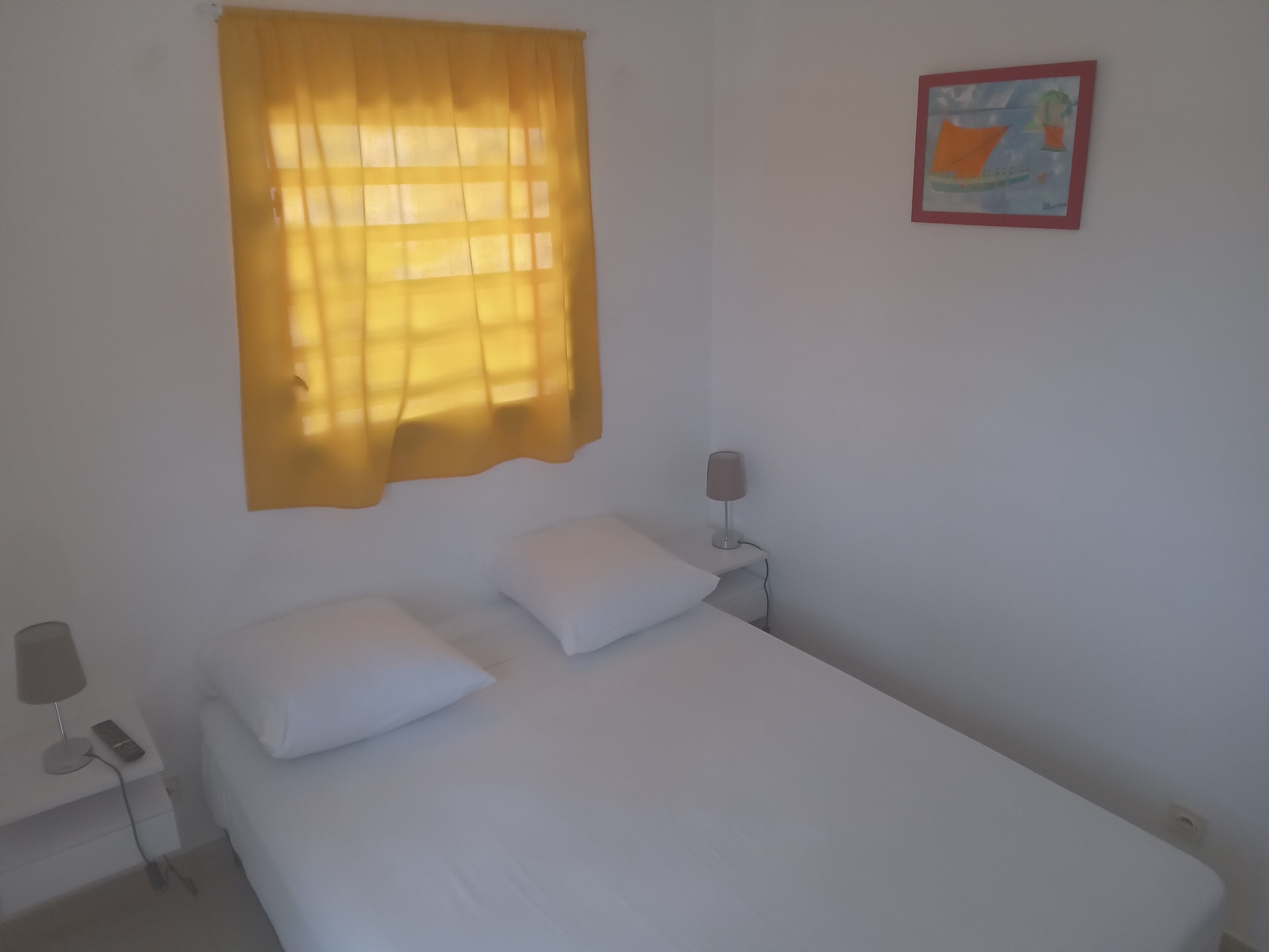 Uma cama de solteiro com roupa de cama branca, uma mesinha de cabeceira com luminária e uma janela com cortinas amarelas.