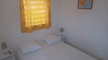 Uma cama de solteiro com roupa de cama branca, uma mesinha de cabeceira com luminária e uma janela com cortinas amarelas.