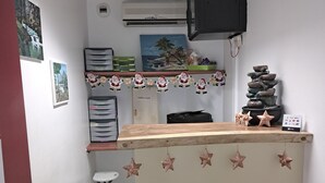 A small reception area with a wooden counter, a small white chair, a desk with a computer, and a wall-mounted TV. There are framed pictures and a small decorative waterfall on the wall. - LE CHANNEL HOTEL (Le Marin)