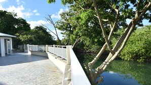 A bridge with a white railing, a paved walkway, and a body of water on one side. - LE CHANNEL HOTEL (Le Marin)