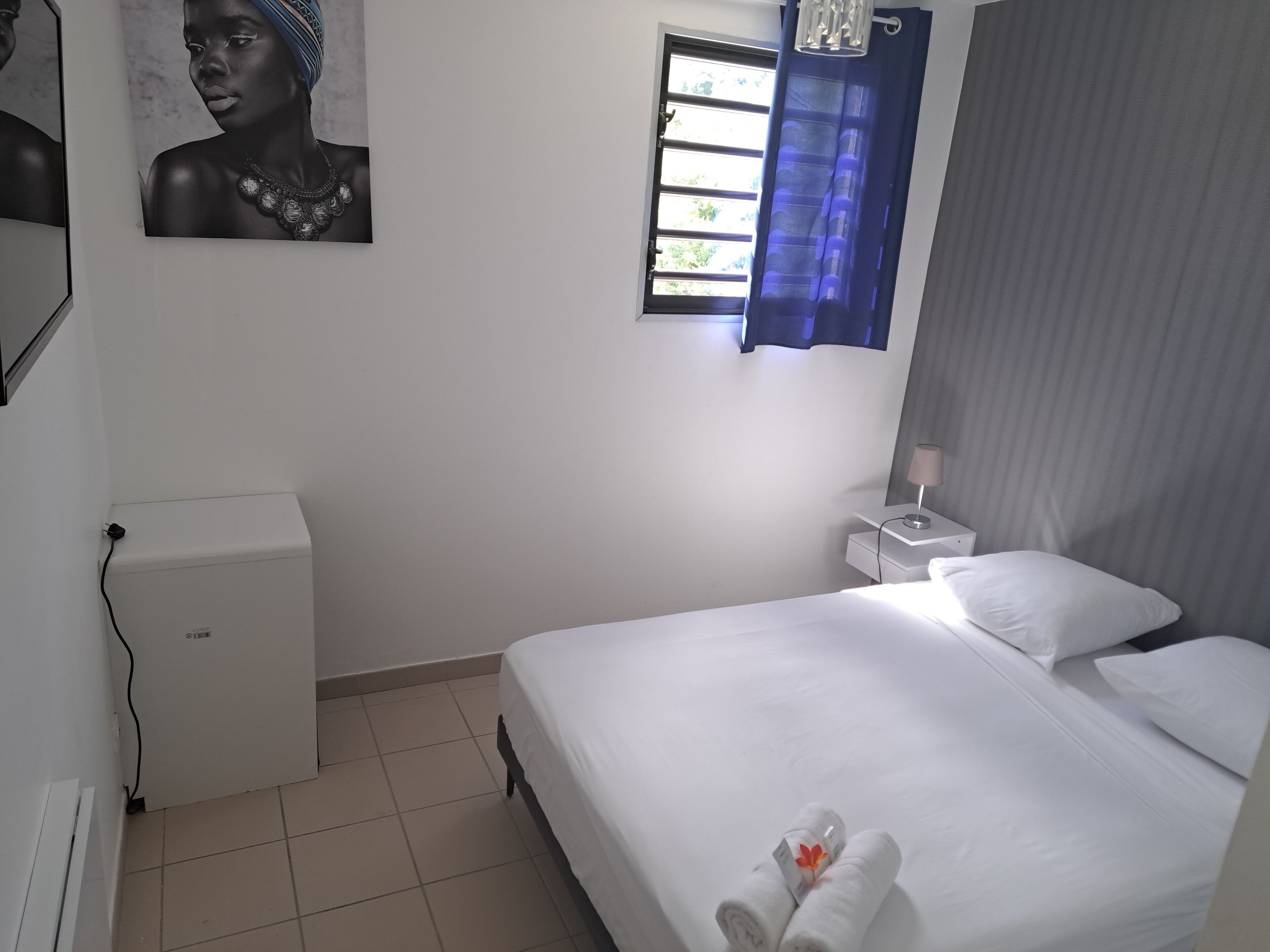 Une chambre d’hôtel avec un lit, une table de chevet, une lampe, une fenêtre avec des rideaux bleus et un tableau accroché au mur.