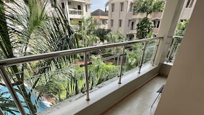 A balcony with a metal railing, a chair, and a view of a pool and greenery. - Cozy & Spacious 3BR Near Beach | Pool & Gym (Dar es Salaam)