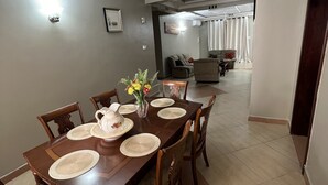 A dining area with a wooden table set for four, surrounded by chairs. A vase with flowers is on the table. - Cozy & Spacious 3BR Near Beach | Pool & Gym (Dar es Salaam)