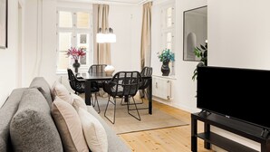 A modern living room with a sofa, a dining table with chairs, a TV, and a large window with curtains.