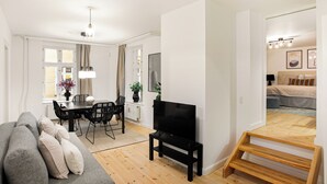 A modern living room with a grey sofa, a dining area with a table and chairs, a TV, and a staircase leading to a bedroom.