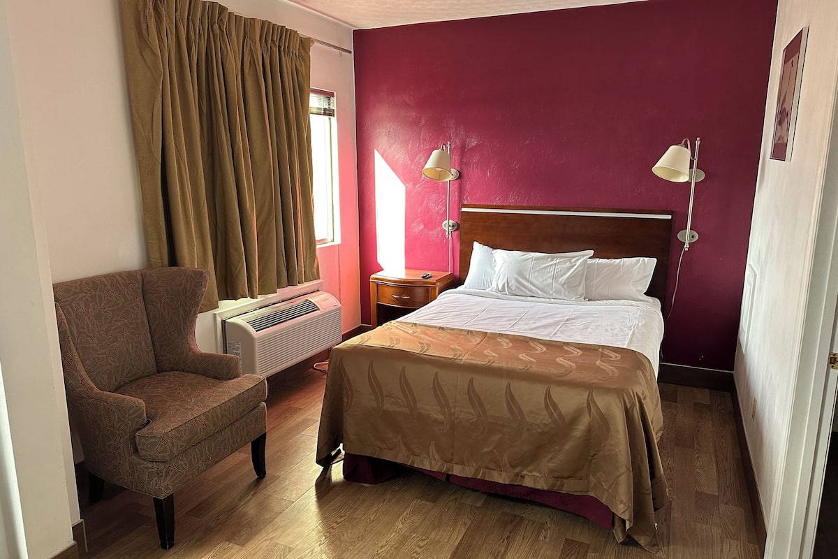 A hotel room with a bed, a chair, a nightstand, a lamp, and a window with curtains.