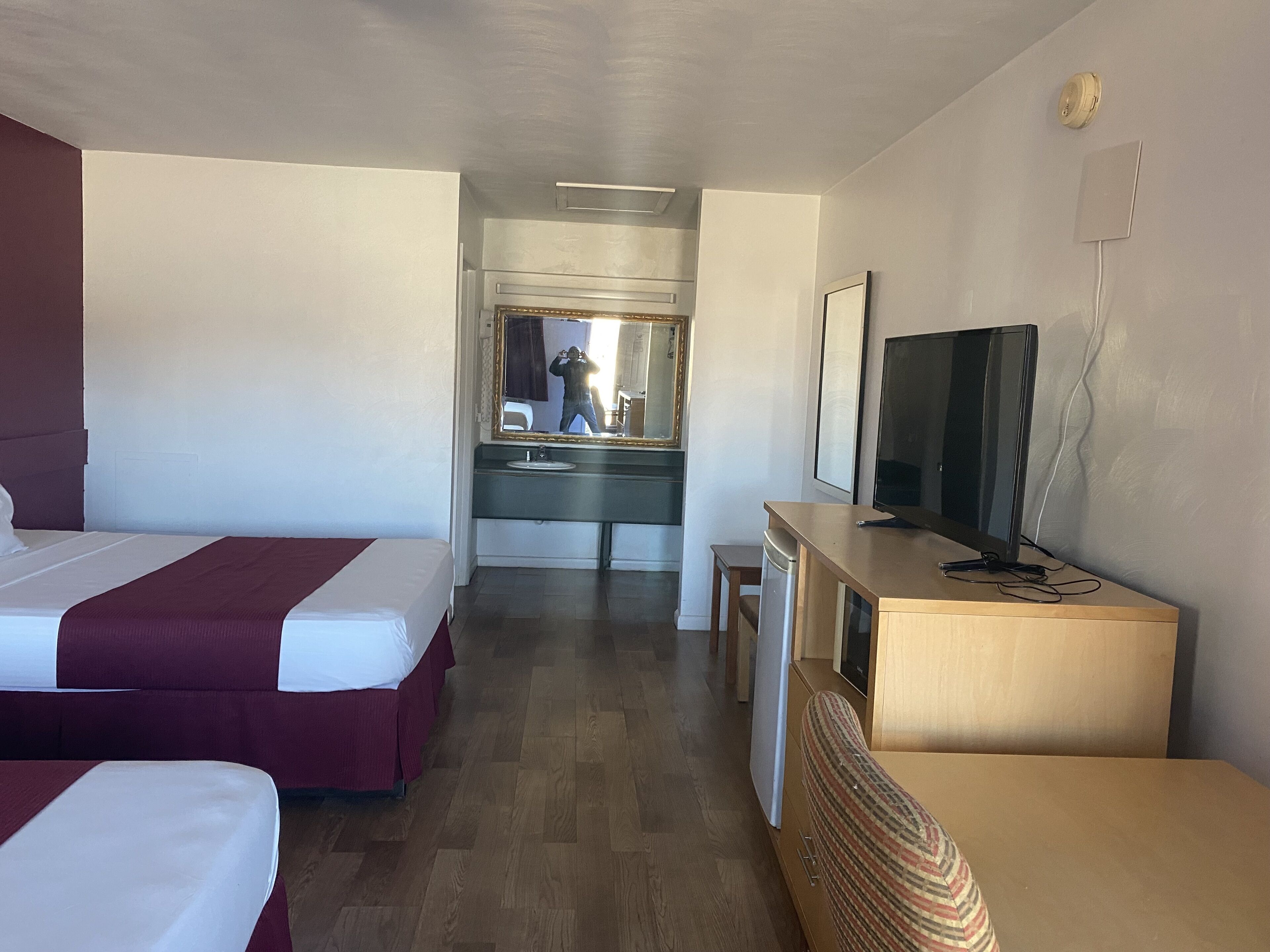 A hotel room with two beds, a television, a desk, a mirror, and a bathroom visible in the background.