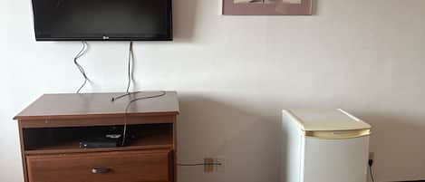 A room with a wooden desk, a flat-screen TV, a small refrigerator, and a framed picture on the wall.