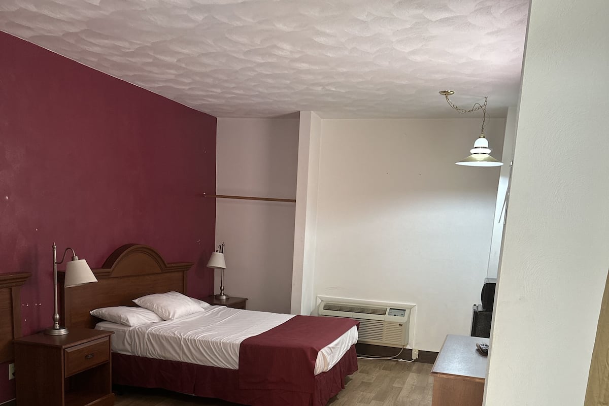A bedroom with a wooden floor, a bed with a red bedspread, a nightstand with a lamp, a heater, and a desk with a chair.