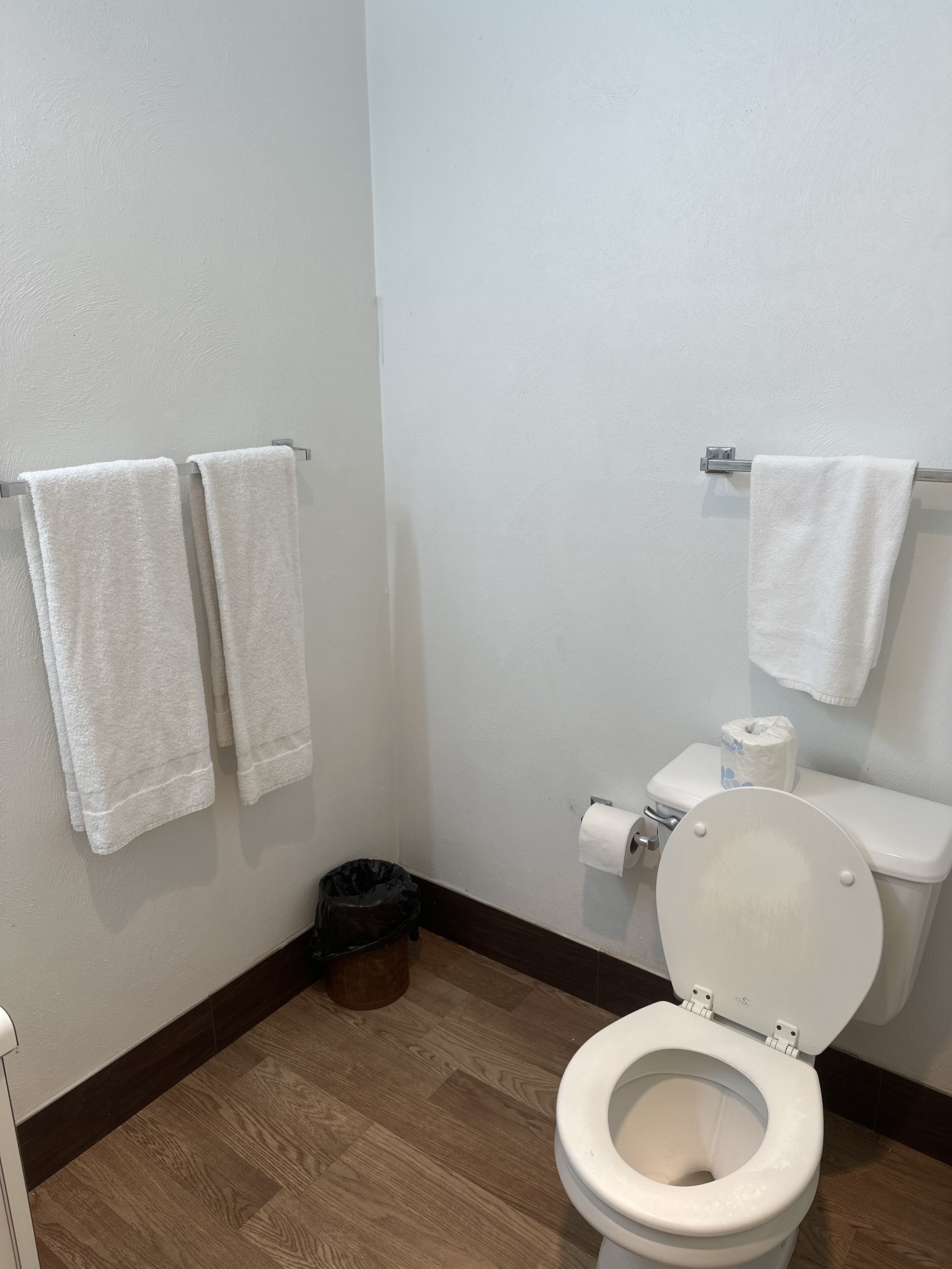 A bathroom with a toilet, two towels on a rack, a roll of toilet paper, and a trash bin.