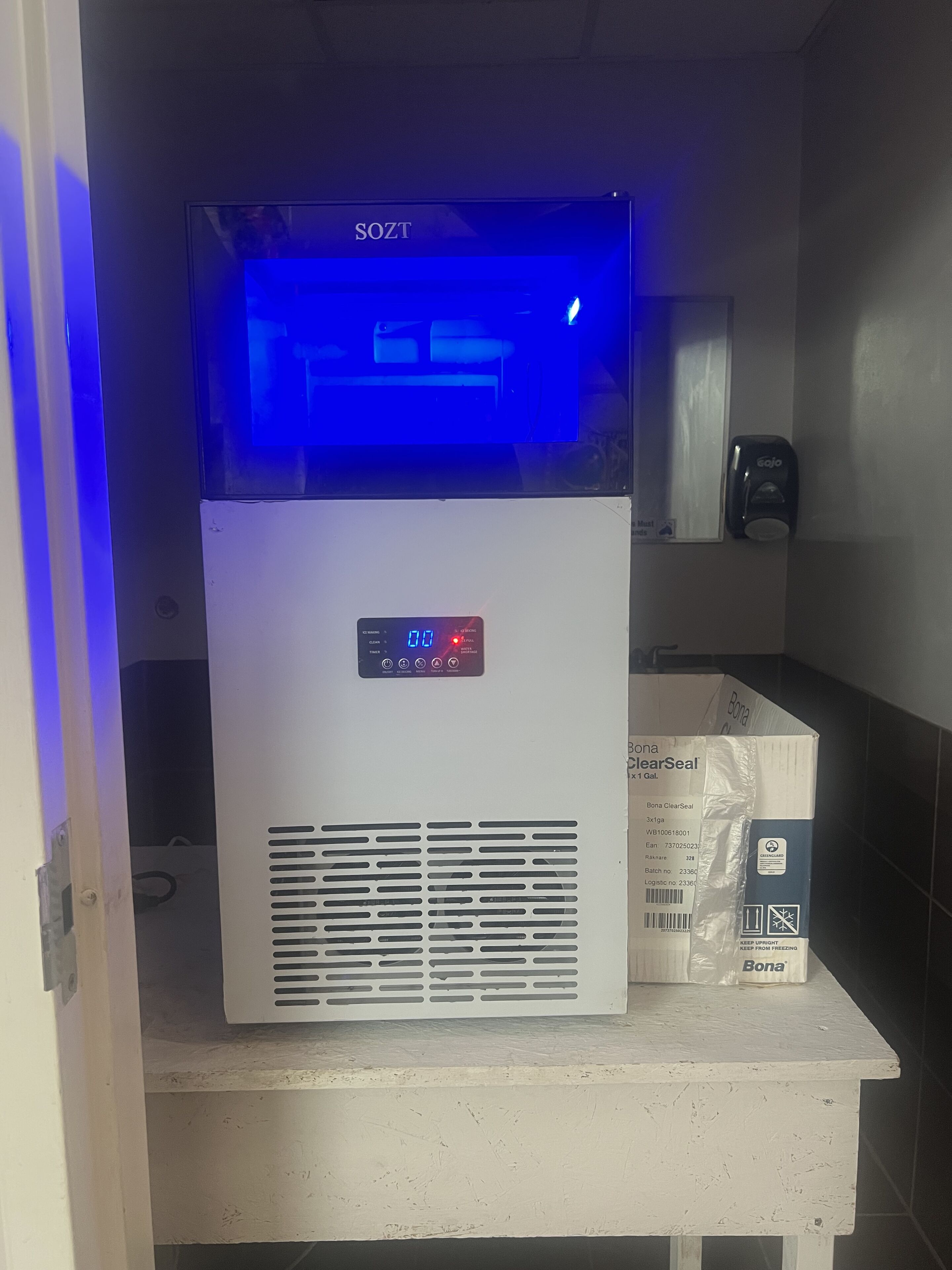 Een SOZT waterdispenser met een digitaal scherm, een witte muur en een BONA ClearSeal doos op een witte toonbank.