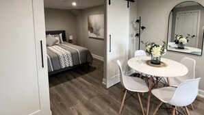 A modern bedroom with a bed, a white dining table with chairs, and a sliding door. - The Chieftain Suite (Barrie)