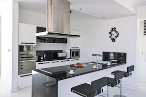 A modern kitchen with a central island, bar stools, stainless steel appliances, and a wall-mounted clock. - KO1 With Breathtaking sea Views (Paralimni)