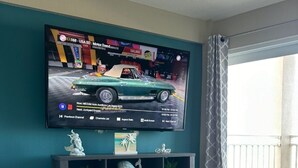 A living room with a flat-screen TV mounted on the wall, a dark wooden entertainment center below, and a grey leather sofa. - Heart of Daytona 1 bedroom w/kitchen, sleeps 4, private balcony, fireplace, pool (Daytona Beach)