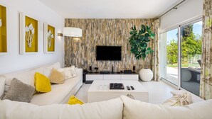 A modern living room with a white sofa, a coffee table, and a flat-screen TV mounted on a textured wall. - Villa Chloe (Vale do Lobo)