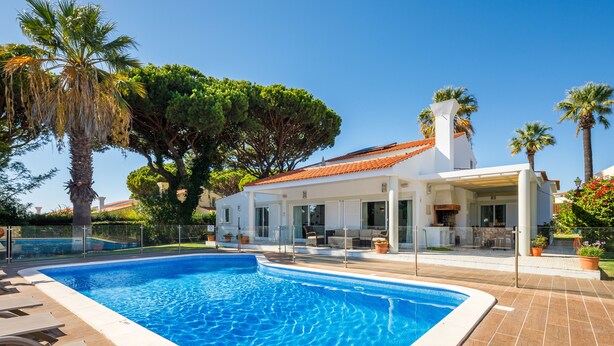 A swimming pool with lounge chairs, a covered patio, and a house with a tiled roof and large windows. - Villa Chloe (Vale do Lobo)