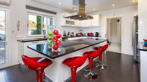 A modern kitchen with a central island, red bar stools, and a glass-top table. - Villa Chloe (Vale do Lobo)