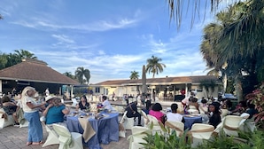 Evento al aire libre con mesas y sillas redondas, palmeras y cielo despejado.