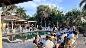Zona de piscina al aire libre con personas sentadas en mesas, rodeada de palmeras y un área de descanso cubierta.