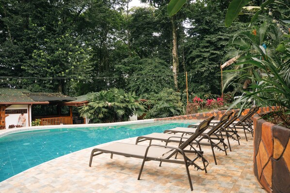 A swimming pool with lounge chairs, surrounded by lush greenery and a wooden deck. - Arenal River Place (La Fortuna)