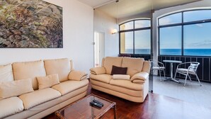 Una sala de estar con sofá, sillón y una mesita de centro. Hay un cuadro en la pared y vistas al mar a través de grandes ventanales.