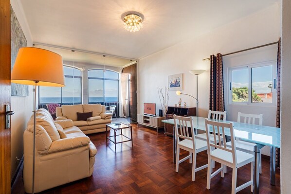 A spacious living area with a sofa set, a dining table with chairs, a TV, and a large window offering a view.