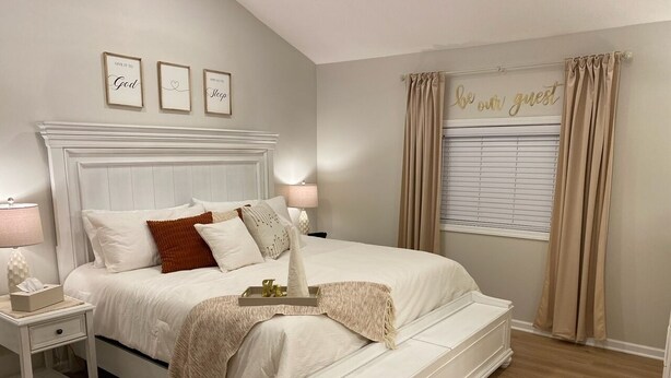 A bedroom with a white bed, beige curtains, and a bedside table with a lamp. - The Landing Zone.
 Comfortable and cozy three bedroom with two car garage.  (East Moline)