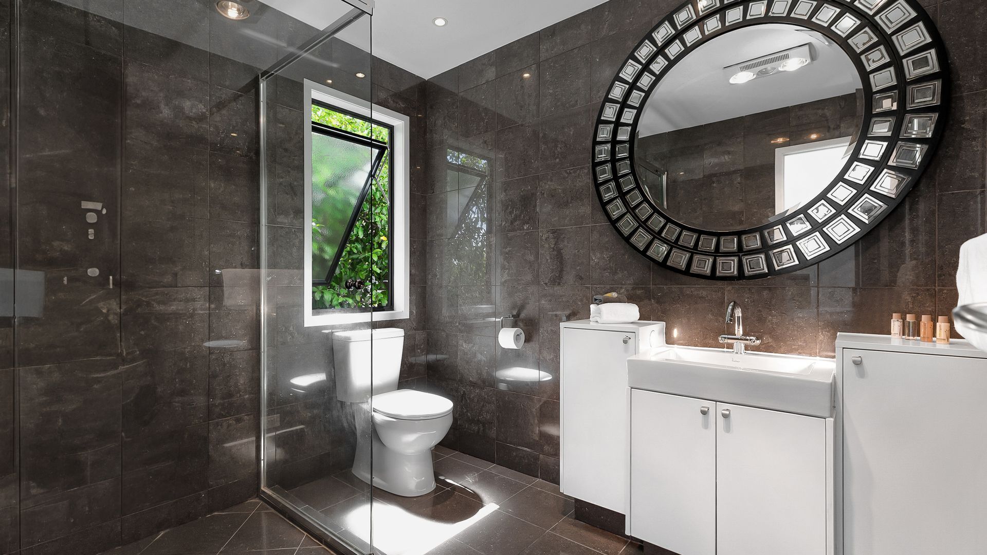 Une salle de bain moderne avec une douche à parois de verre, des toilettes, un lavabo avec un miroir et une fenêtre donnant sur un espace verdoyant.