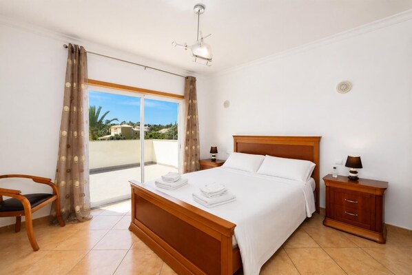 A hotel room with a bed, a chair, a nightstand, a lamp, and a view of the outside.