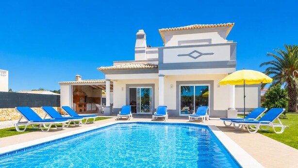 A swimming pool with blue water, surrounded by lounge chairs and a building with a tiled roof.