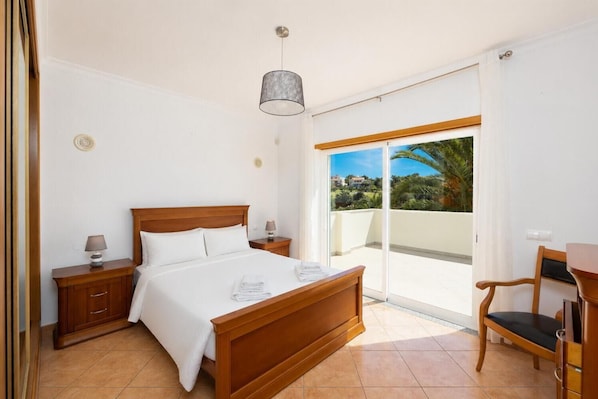 Un dormitorio con una cama de madera, mesitas de noche, una lámpara, una silla y vistas a un balcón con una palmera.