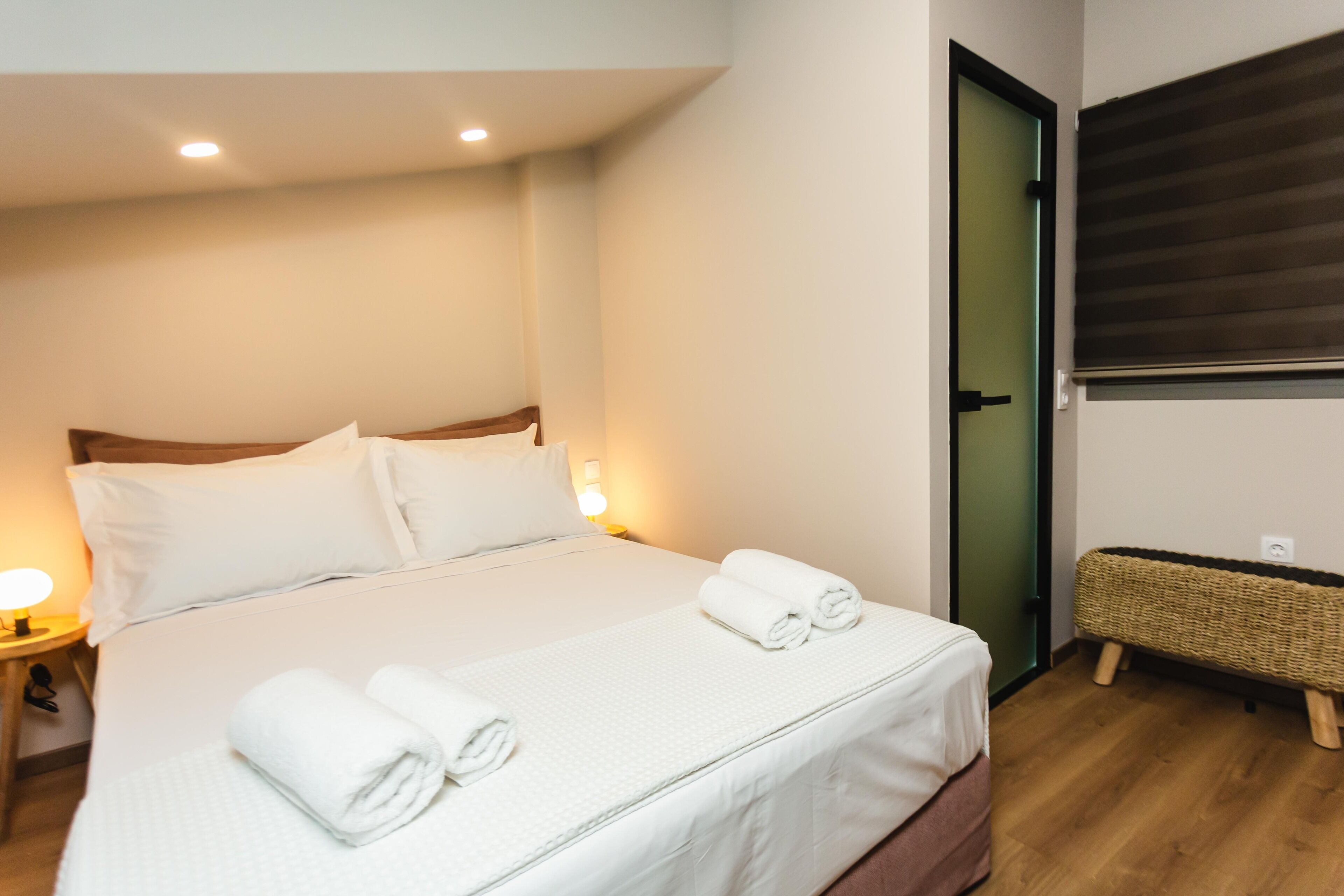 A neatly made bed with white linens and rolled towels, a bedside lamp, and a wicker bench against a wall.