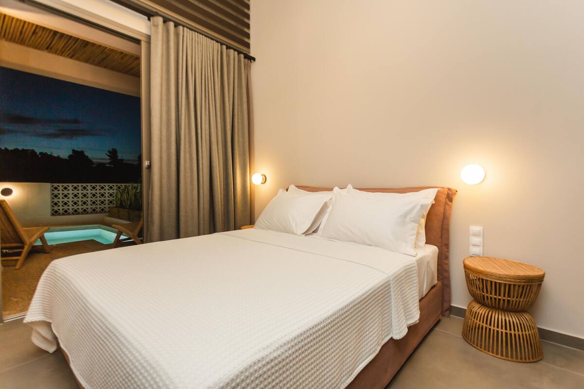 A bedroom with a large bed, two bedside lamps, a wicker side table, and a view of the outdoors.