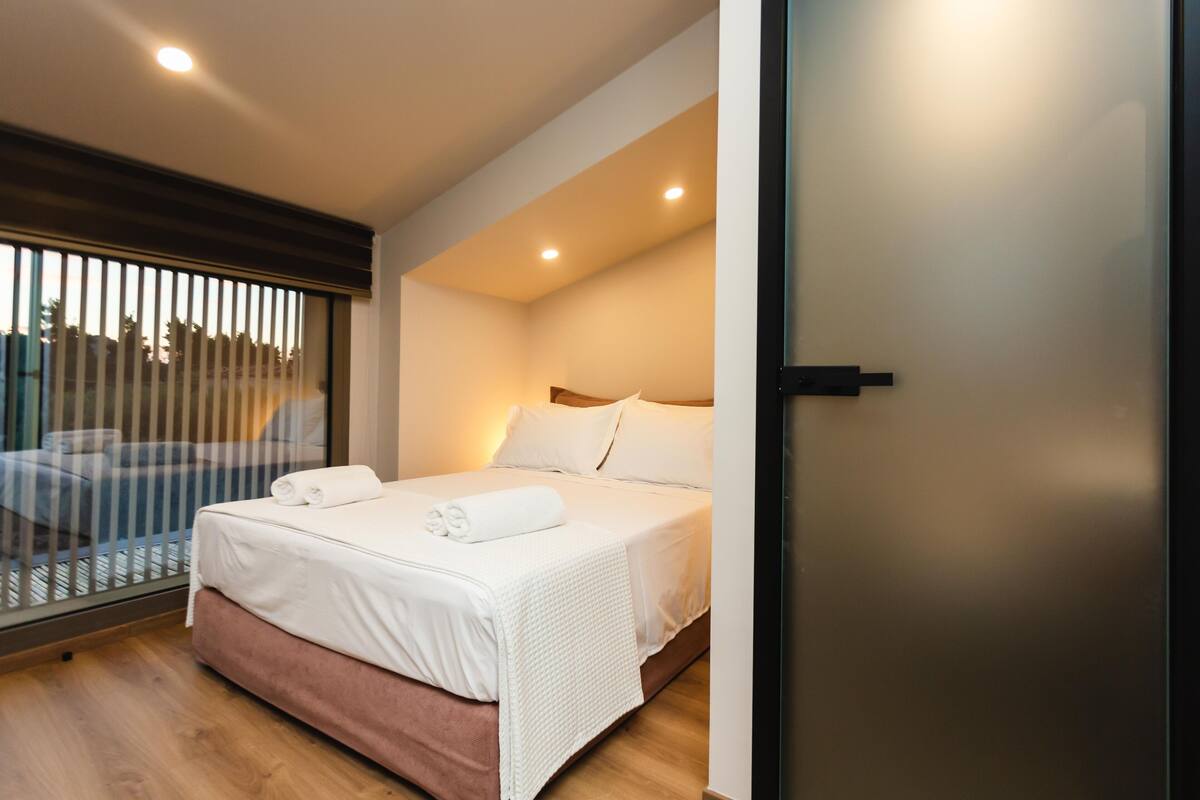 A modern hotel room with a large bed, a window with blinds, and a wooden floor.