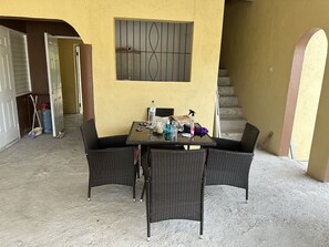 Un coin repas avec une table et quatre chaises en osier, une fenêtre avec des barreaux et un escalier en arrière-plan.