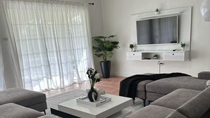 Un salon moderne avec un canapé d’angle gris, une table basse agrémentée d’objets décoratifs, une grande plante en pot et un téléviseur à écran plat fixé au mur.