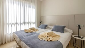 Habitación de hotel con dos camas, un ventanal con cortinas translúcidas, una mesita de noche con lámpara y un interruptor de luz en la pared.
