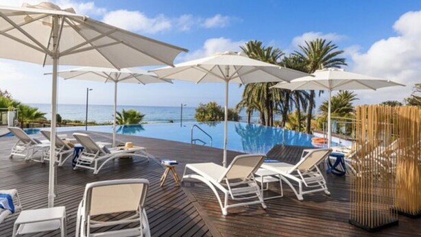 Zona junto a la piscina con sillones y sombrillas, con vista a un cielo azul claro y al océano.